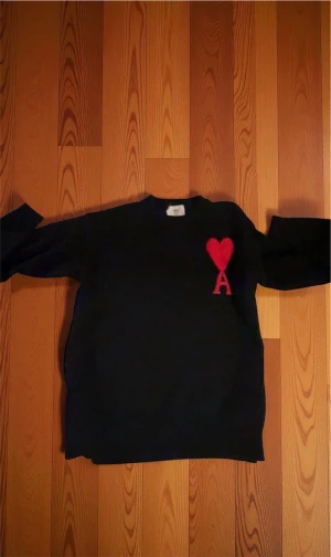 Svart stickad tröja från Ami med hjärta - Svart stickad tröja från Ami med ett stort rött hjärta och bokstaven A broderat på bröstet. Rund hals och ribbade muddar vid ärmslut och nederkant. Perfekt för dig som gillar stilrena detaljer och vill sticka ut med en ikonisk design.