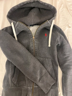 Grå hoodie från Polo Ralph Lauren - Mysig grå hoodie från Polo Ralph Lauren med vit snörning i huvan och röd logga på! Denna är bra skick men har lite slitningar men inte sönder 