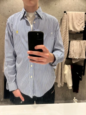 Blåvit randig skjorta från Ralph Lauren - Snygg klassisk skjorta från Ralph Lauren i blått och vitt med smala vertikala ränder. Classic fit med button-down krage och gul broderad logga på bröstet. Skjortan har långa ärmar och vita knappar framtill. Perfekt för dig som gillar stilrena och tidlösa plagg.