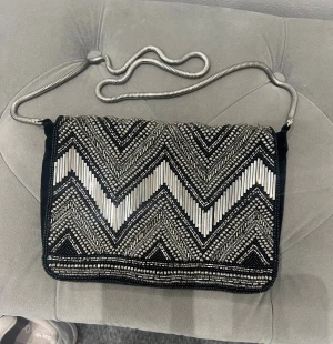 Silvrig kuvertväska med zigzag-mönster - Snygg kuvertväska med glittrigt zigzag-mönster i silver och svart. Väskan har en metallkedja som axelrem och är täckt av pärlor och paljetter för extra bling. Perfekt för att lyfta din outfit med lite edge och glam.
