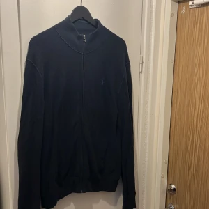 Polo Ralph Lauren marinblå kofta  - Säljer en stilren marinblå kofta från Polo Ralph Lauren med hel dragkedja framtill och diskret ljusblå logga på bröstet. Tröjan är stickad i bomull och har en hög krage samt långa ärmar. Perfekt för lager-på-lager och snygg till jeans.
