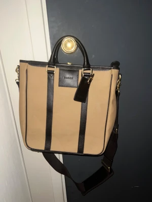 Datorväska från Baron - Snygg beige datorväska från Baron med mörkbruna skinn-detaljer och guldfärgade metalldelar. Väskan har både handtag och justerbar axelrem, perfekt för att bära med sig allt viktigt. Klassisk och stilren design med rektangulär form. Aldrig använd, dustbag medföljer