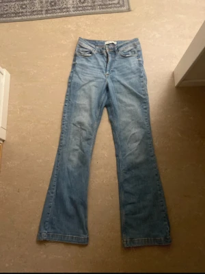 Bootcut ljusblå jeans med rå kant - Säljer ett par ljusblå bootcut jeans med klassisk femficksdesign och rå, fransig kant längst ner på benen. Jeansen har hög midja och är tillverkade i mjukt denimtyg. Perfekt för dig som gillar en avslappnad och trendig look.