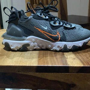 Nike React Element sneakers grå/orange - Nike React Element sneakers i grått med svarta och orange detaljer. Snygg design med vit och svart mönstrad sula, svart snörning och ventilerande ovandel i syntet. Ikonisk orange swoosh på sidan och coola detaljer på hälen. Perfekta för dig som gillar streetstyle.