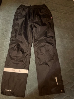 Svarta  skalbyxor från Tretorn Teknees - Svarta skalbyxor från Tretorn Teknees med elastisk midja och snörning. Byxorna har reflexdetalj på ena benet, knappstängning vid benslut och praktiska fickor. Perfekta för kalla och blöta dagar utomhus.