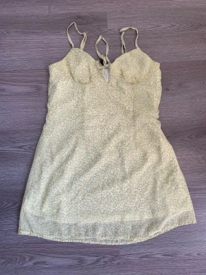 Blommig ljusgul kort sommarklänning - Supersöt ljusgul kort klänning med småblommigt mönster i grönt och gult från H&M. Klänningen har smala axelband som knyts och en lätt figurnära passform. Perfekt för varma dagar och ger en fräsch och romantisk vibe.