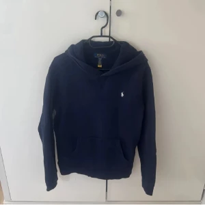 Mörkblå hoodie från Polo Ralph Lauren - Snygg mörkblå hoodie från Polo Ralph Lauren med klassisk huva och känguruficka framtill. Tröjan har lång ärm och den ikoniska vita Polo-loggan broderad på bröstet. Perfekt för chill dagar och enkel att matcha med jeans eller joggers.