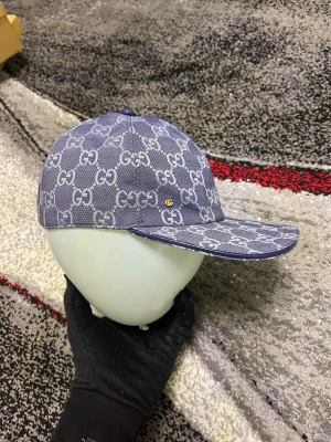 Blå Gucci keps med GG-mönster - Snygg blå keps från Gucci med ikoniskt GG-mönster över hela kepsen. Kepsen har en klassisk form med böjd skärm och justerbar rem baktill. Materialet är bomull och insidan är ljusbeige. Perfekt för dig som vill ha en lyxig streetstyle-accessoar.