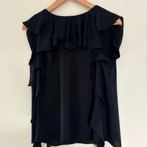 Svart blus med volanger från Tara Jarmon - Supersnygg svart blus från Tara Jarmon i storlek 38. Blusen är ärmlös och har volanger längs axlar och halsringning samt en rosett i nacken. Materialet är lätt och flowy, perfekt för dig som gillar en chic och feminin stil.