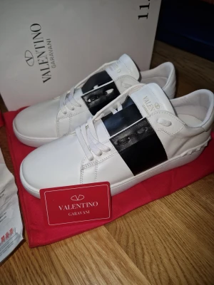 Valentino Garavani Open sneakers vit/svart - Snygga Valentino Garavani Open sneakers i vitt skinn med bred svart läderdetalj över sidan. Klassisk rund tå, platt sula och vita snören. Märkeslogga i guld på plösen. Perfekt för dig som gillar stilrena och lyxiga sneakers med unik design.