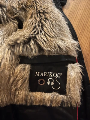 Svart vinterjacka med fuskpäls från Marikoo - Säljer en svart vinterjacka från Marikoo i storlek S. Jackan har ett quiltat innerfoder och är fodrad med mjuk, beige fuskpäls på insidan och i huvan. Materialet är polyester och jackan har snygga detaljer som logga och hörlursuttag.