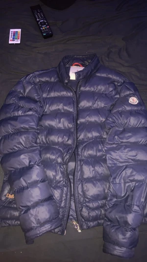 Mörkblå dunjacka från Moncler - Säljer en mörkblå Moncler dunjacka med klassisk quiltad design och dragkedja framtill. Jackan har hög krage, Moncler-logga på ärmen och coolt serietidningsfoder på insidan. Perfekt för kalla dagar och riktigt snygg streetstil.