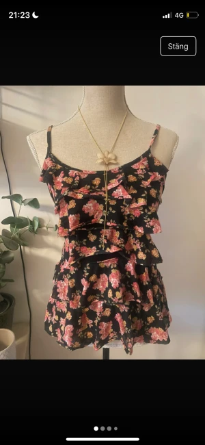 Svart blommig volangklänning - Supersöt kort klänning med svart botten och blommigt mönster i rosa och gult. Klänningen har smala axelband, flera volanger framtill och figurnära passform. Perfekt för dig som gillar romantisk och lekfull stil.