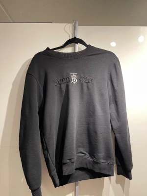 Svart sweatshirt från Burberry - Svart sweatshirt från Burberry med broderad logga och klassiskt TB-märke på bröstet. Tröjan har rund hals, ribbade muddar och lång ärm. Mjuk bomullskvalitet som är skön att bära och passar perfekt till jeans eller cargo.