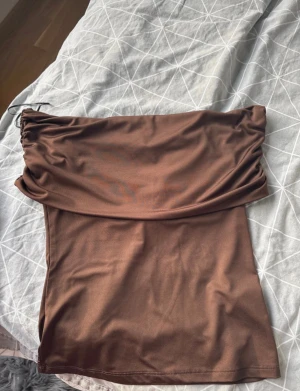 Offshoulder tröja från  H&M - Snygg brun off shoulder tröja från H&M i storlek M. Tröjan har en cool snörningsdetalj i midjan och är gjord i mjukt, stretchigt syntetmaterial. Perfekt för dig som gillar minimalistisk stil med en twist. Passformen är normal och tröjan har en enkel, rak siluett.