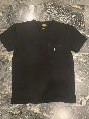 Svart t-shirt från Polo Ralph Lauren - Svart t-shirt från Polo Ralph Lauren med klassisk passform och korta ärmar. T-shirten har en bröstficka med den ikoniska vita Polo-loggan broderad. Tillverkad i mjuk bomull som känns skön mot huden. Perfekt för en clean och stilren look. 