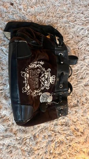 Juicy Couture brun axelväska med tryck - Snygg brun axelväska från Juicy Couture med svart skinn och mocka, detaljerad med metallplakett 'Royal Juicy' och hjärtformad charm. Väskan har ett broderat vitt tryck på framsidan, snörning upptill och insida med silverfärgad fack och tryckt text. Osäker om den är äkta men färgen på insidan har lossnat men går nog att fixa, inga synliga defekter från utsidan 