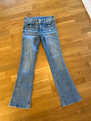  Nya Bootcut jeans från H&M ljusblå - Ljusblå bootcut jeans från H&M med låg midja och justerbar midja. Klassisk femficksmodell med två framfickor med lock och knappar. Jeansen har en snygg tvätt och dubbla knappar i midjan. Perfekta för dig som gillar retrovibbar och bekväm passform.