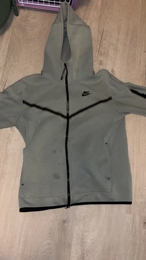 Grå Nike tech hoodiejacka med dragkedja - Grå hoodiejacka från Nike med hel dragkedja och svart Nike-logga på bröstet. Jackan har svarta detaljer längs dragkedjan och över bröstet, samt fickor på sidorna. Tillverkad i mjukt syntetmaterial och har lång ärm och huva.