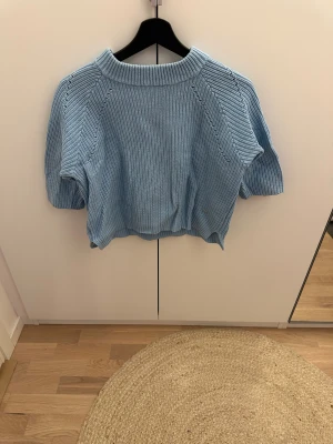 Ljusblå stickad tröja Holly & Whyte - Snygg ljusblå stickad tröja från Holly & Whyte med korta ärmar och ribbad struktur. Tröjan har rund halsringning och en liten metallbricka med logga framtill. Perfekt för dig som gillar chill och stilren vibe.