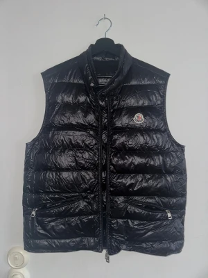 Moncler gui vest svart - Svart quiltad dunväst från Moncler med glansig finish och ståkrage. Västen har två fickor med dragkedja framtill och klassisk Moncler-logga på bröstet. Modellen är Longue Saison och har en stilren, normal passform. Perfekt för lager-på-lager under kyliga dagar.