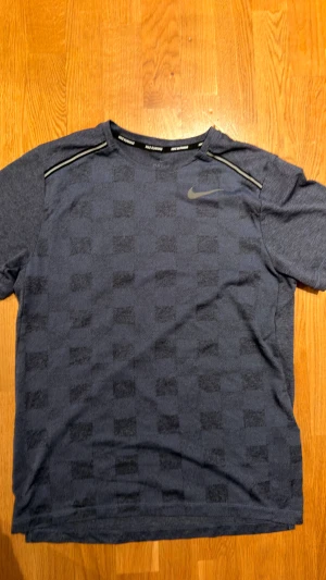 Blå Nike Dri-Fit t-shirt med mönster - Säljer en blå Nike Dri-Fit t-shirt med diskret rutmönster och reflekterande detaljer vid axlarna. Perfekt för löpning eller träning, tillverkad i ett lätt och snabbtorkande material. Klassisk Nike-logga på bröstet och rund hals.