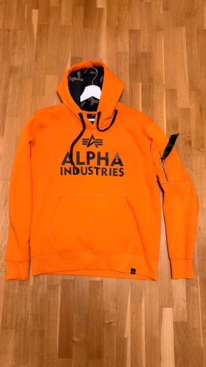 Orange hoodie från Alpha Industries - Snygg orange hoodie från Alpha Industries med svart logotryck på bröstet och cool detalj med dragkedja på ärmen. Hoodien har snörning i huvan och en klassisk känguruficka framtill. Insidan av huvan har ett camouflagemönster för extra stil.