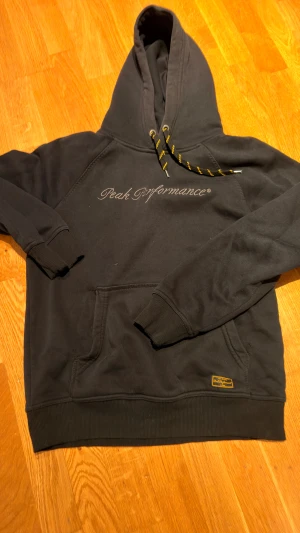 Svart hoodie Peak Performance M - Svart hoodie från Peak Performance med broderad logga på bröstet och coola svarta snören med gula detaljer. Tröjan har huva, känguruficka och ribbade muddar. Mjuk insida i fleece som gör den extra skön. Perfekt för chill eller häng med kompisar.