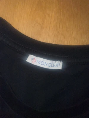 Svart Moncler t-shirt med ränder - Snygg svart t-shirt från Moncler med klassisk logga på bröstet och vita ränder längs axlarna. T-shirten har rund hals och korta ärmar, perfekt för en clean och sportig look. Tillverkad i mjuk bomull för skön känsla hela dagen. Frågor kan skrivas till mig. Har bevis på att den är äkta.