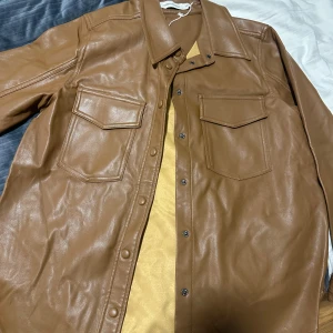 Brun skinn overshirt från Carin Wester - Säljer en brun overshirt i skinn från Carin Wester. Jackan har två stora bröstfickor med lock, tryckknappar framtill och klassisk krage. Perfekt för dig som vill ha en stilren och trendig look med en oversized känsla.