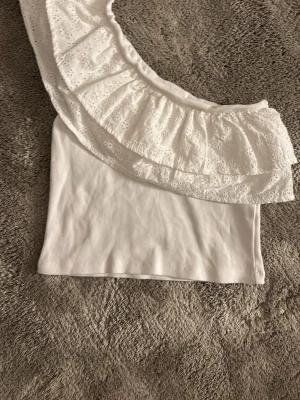 Fint sommarlinne  - Superfin vit offshoulder topp från Gina Tricot med bred volang och broderade spetsdetaljer. Toppen är ribbad och har en ärmlös, asymmetrisk design som ger en trendig look. Perfekt att styla till jeans eller kjol för en fräsch sommaroutfit.