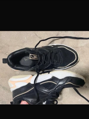 Svarta sneakers från Puma - Säljer ett par chunky svarta sneakers från Puma med vita och beige detaljer. Skorna har snörning och en tjock, vit sula som ger en sportig vibe. Materialet är en mix av skinn och syntet, vilket gör dem både snygga och bekväma till vardags.