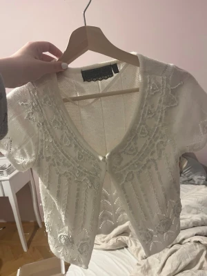 Vintage Vit stickad kofta med paljetter - Supersöt vit stickad kofta från Vintage Stories med korta ärmar och v-ringning. Koftan har fina paljett- och pärldetaljer framtill och stängs med små hyskor. Perfekt för att lyfta en outfit med lite extra glitter.