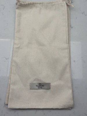 Beige dustbag från Valentino Garavani 2 st - Säljer en beige dustbag från Valentino Garavani i bomull med dragsko upptill och en snygg logotypetikett framtill. Perfekt för att förvara din väska eller accessoarer på ett stilrent sätt. 2 st