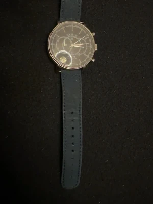 Mörkblå GANT klocka med läderband ( Gant Cameron W10891)  - Snygg klocka från GANT med mörkblått läderband och silverfärgat boett. Urtavlan har flera små visare och detaljer, vilket ger en modern och stilren look. Klockan har klassisk rund form och diskreta knappar på sidan.