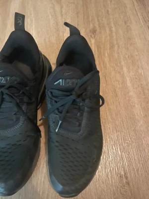 Svarta Nike Air Max 270 sneakers - Säljer ett par helsvarta Nike Air Max 270 sneakers med mesh-överdel och synlig luftbubbla i hälen. Skorna har snörning, rund tå och en sportig vibe. Perfekta för dig som gillar stilrena och bekväma sneakers med modern design.
