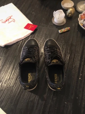 Svarta Fendi sneakers med FF-mönster - Snygga svarta sneakers från Fendi med klassiskt FF-mönster i mörkgrått och svart. Skorna har vita sulor, svarta skinnpartier vid tå och häl samt svarta snören. Fendi-logga i gult på plösen och insidan. Perfekta för dig som vill sticka ut med lyxig streetstyle.