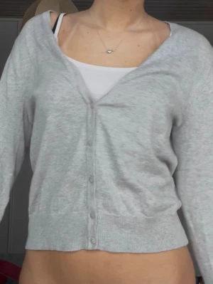 Ljusgrå kofta med knappar - Ljusgrå kofta med v-ringning och knäppning framtill. Tröjan har långa ärmar och ribbade muddar vid ärmslut och nederkant. Perfekt att slänga över ett linne eller t-shirt för en chill och enkel look.