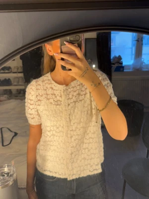 Vit blus från Motel - Supersöt vit blus från Motel i spets med blommigt mönster. Kort ärm och knappar hela vägen framtill. Perfekt att styla över ett linne för en romantisk vibe. Lätt och luftig, passar perfekt till vår och sommar.