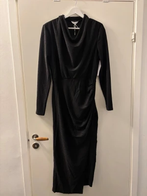 Svart glittrig långklänning Bubbleroom - Säljer en svart långklänning från Bubbleroom med glittrigt tyg och draperad halsringning. Klänningen har långa ärmar och snygga rynkade detaljer längs midjan och höften. Perfekt för dig som vill ha en elegant look med lite extra shimmer. ALDRIG ANVÄNT etiketten finns kvar