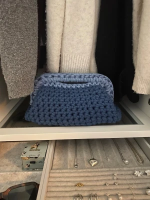Blå handgjord stickad handväska - Säljer en superfin handväska i virkad design, gjord i ljusblått/mörkblått second hand t-SHIRT garn från massindustrin. Väskan har en mjuk, rektangulär form och ett praktiskt handtag upptill (20cm). Perfekt för att bära småsaker i och ger en unik touch till din outfit, oavsett om du ska till skolan eller ut på stan!