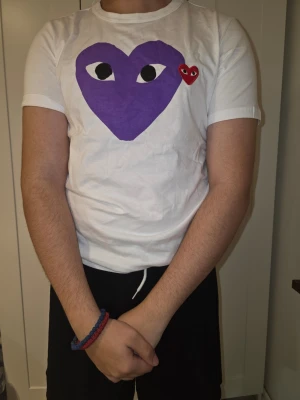 Vit Comme des Garçons Play t-shirt - Säljer en vit Comme des Garçons Play t-shirt med det ikoniska hjärtat i lila och ett mindre rött hjärta på bröstet. Klassisk rund hals och korta ärmar. Tillverkad i mjuk bomull och har en clean, stilren look som sticker ut.