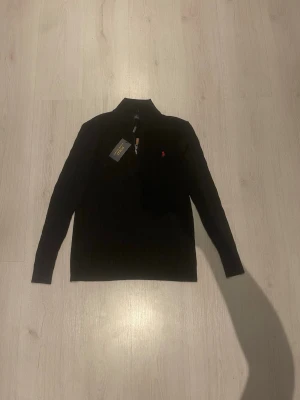 Svart stickad tröja Polo Ralph Lauren XL - Snabb leverans. Snygg svart stickad tröja från Polo Ralph Lauren i storlek XL. Tröjan har kabelstickat mönster, hög krage och half zip-dragkedja med läderdetalj. Klassisk röd logga broderad på bröstet. Perfekt till höst och vinter! Det står XL men känns som M
