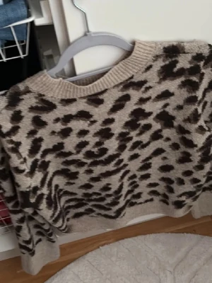 Beige leopardmönstrad stickad tröja H&M - Supermysig stickad tröja från H&M i beige med brunt leopardmönster. Tröjan har rund halsringning, ribbade muddar och är långärmad. Perfekt för att lyfta din höst- eller vinterstil med lite edge.