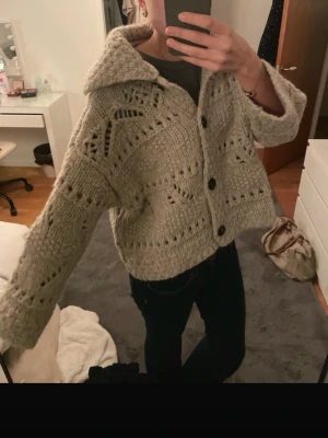 Grå stickad kofta från Zara - Säljer en  grå stickad kofta från Zara med snyggt hålmönster och stora svarta knappar framtill. Koftan har en bred krage och lång ärm, perfekt för lager på lager under kyliga dagar.