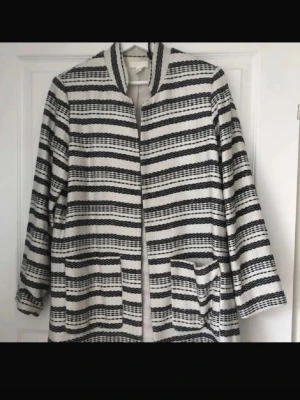 Randig svartvit kappa med fickor - Snygg kappa i svartvitt randigt mönster med raka linjer och två stora fickor framtill. Jackan har en öppen design utan synliga knappar eller dragkedja och en ståkrage. Perfekt för dig som gillar stilrena och grafiska plagg.