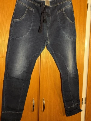 Please jeans Mörkblå jeansbyxor från  - Mörkblå jeansbyxor från Please med bekväm resårmidja och svart snörning. Byxorna har stora framfickor, två bakfickor och snygga sömdetaljer. Materialet är stretchigt jeans och passformen är loose med avsmalnande ben.
