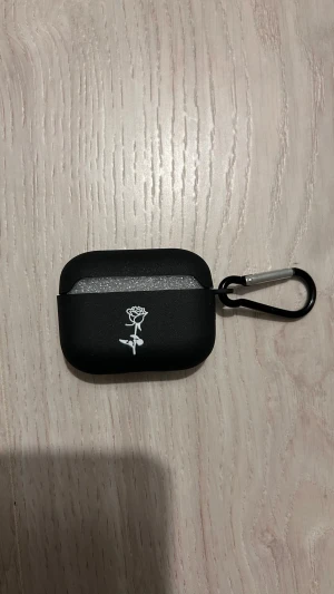 AirPods Pro (2nd gen) med svart fodral och karbinhake - Snygga AirPods Pro (2nd gen) i svart skyddsfodral med vit rosdesign och karbinhake för enkel upphängning. Fodralet ser ut att vara i mycket gott skick utan synligt slitage. Perfekt för dig som vill ha trådlösa hörlurar med stil och funktion.