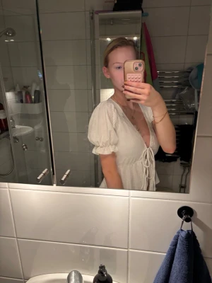 Vit blus med knyt och puffärm - Supersöt vit blus med djup v-ringning, puffiga korta ärmar och knyt framtill. Toppen har en lätt strukturerad bomullskänsla och sitter lite lösare nedtill. Perfekt för en fräsch och somrig look.