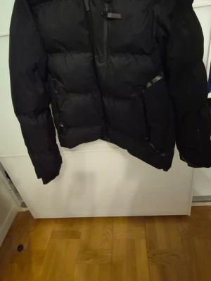 Svart pufferjacka från Jack & Jones - Säljer en svart pufferjacka från Jack & Jones Core. Jackan har dragkedja framtill, stora fickor och justerbar huva. Perfekt för kalla dagar och har en clean, modern look. Passar dig som gillar streetwear och vill hålla värmen.